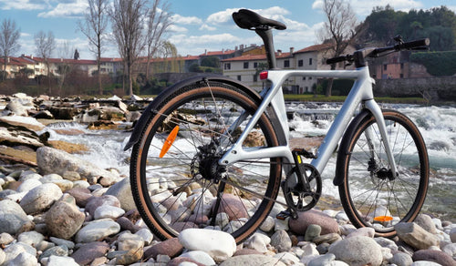 Ultralekki rower elektryczny Fiido Air z włókna węglowego, prezentowany na kamienistym brzegu rzeki obok płynącej wody.