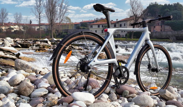 Ultralekki rower elektryczny Fiido Air z włókna węglowego, prezentowany na kamienistym brzegu rzeki obok płynącej wody.