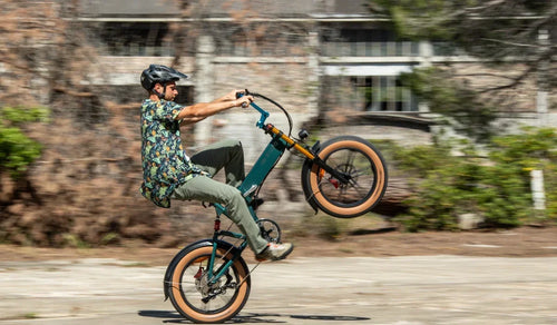 Rowerzysta jedzie na tylnym kole na elektrycznym fat bike’u Fiido M1 Pro, prezentując na miejskiej drodze jego kompaktową, składaną ramę, szerokie opony i przednie zawieszenie.
