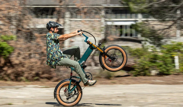 Rowerzysta jedzie na tylnym kole na elektrycznym fat bike’u Fiido M1 Pro, prezentując na miejskiej drodze jego kompaktową, składaną ramę, szerokie opony i przednie zawieszenie.